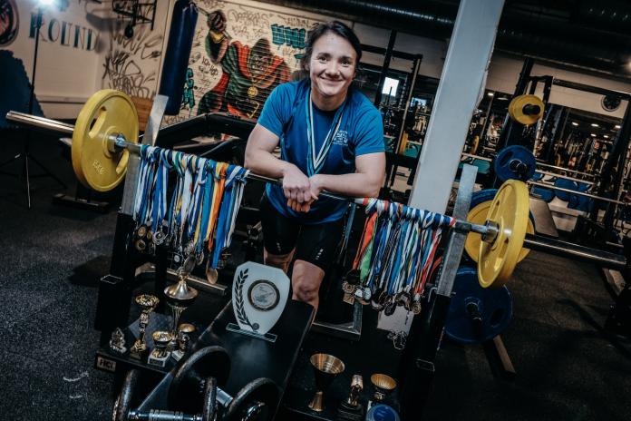 A female Para powerlifter poses behind the barbell from which numerous medals hang.