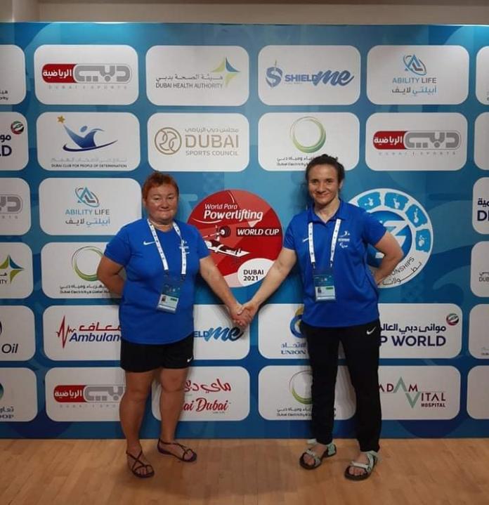 Two women hold hands as they pose next to the official backdrop of the Dubai 2021 World Para Powerlifting World Cup.