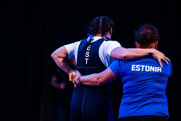 A back view of two women, an athlete and a coach, as they leave the competition field embracing each other.