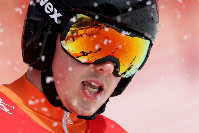 A close up of a male skier's face in goggles as snowflakes fly around him.