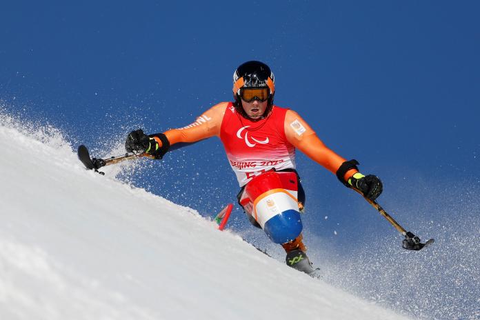 A male sit skier brushes past a slalom pole on a steep alpine slope as snow flies out from his outriggers.