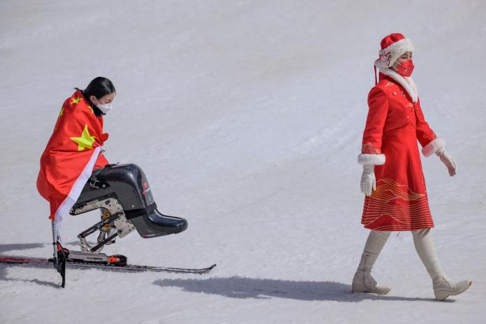 A female sit skier draped in China's flag follows a woman wearing a red dress.