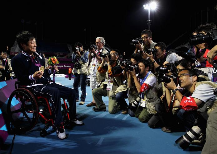 A male wheechair tennis athlete holds a gold medal in front of about 10 photographers taking photos of him