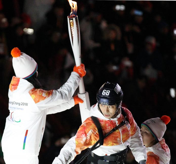 A woman attaches the Paralympic torch onto equipment that a man is carrying on his back