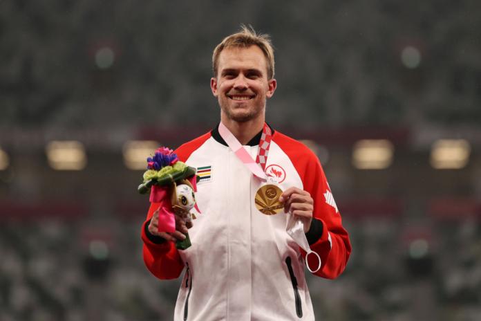 A male athlete smiles while holding a gold medal with his left hand.