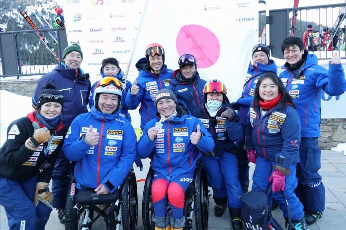 11 athletes and staff of the Japanese Para alpine skiing team pose for a photograph in front of the Japanese flag.
