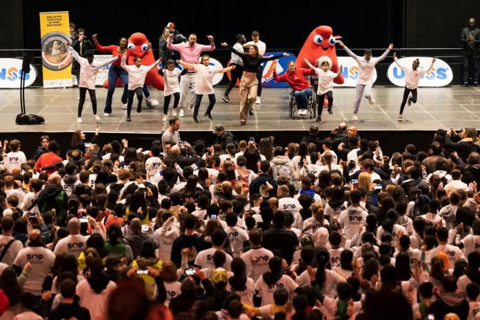 About 15 people dance on a stage with Paris 2024 Games mascots in front of many people