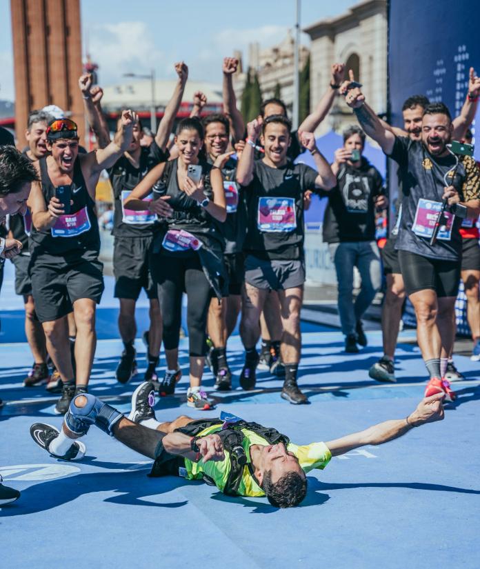 A male runner lies down at the finish line of a marathon, his hands in the air as his supporters run smiling towards him.