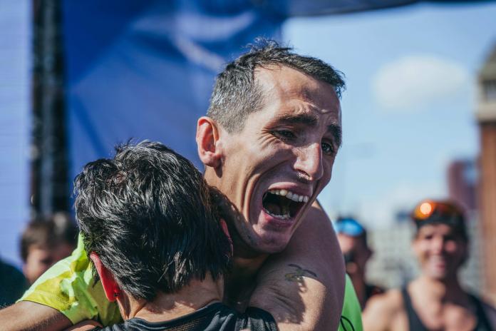 A male runner gasps in joy as he is hugged at the finish line of a marathon.