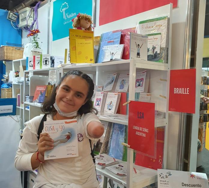 A young girl who is missing her left hand stands by a bookshelf and holds up a book about Paralympic swimmer Daniela Giménez.