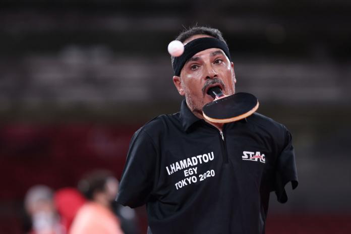 A male athlete plays table tennis with the racquet in his mouth.