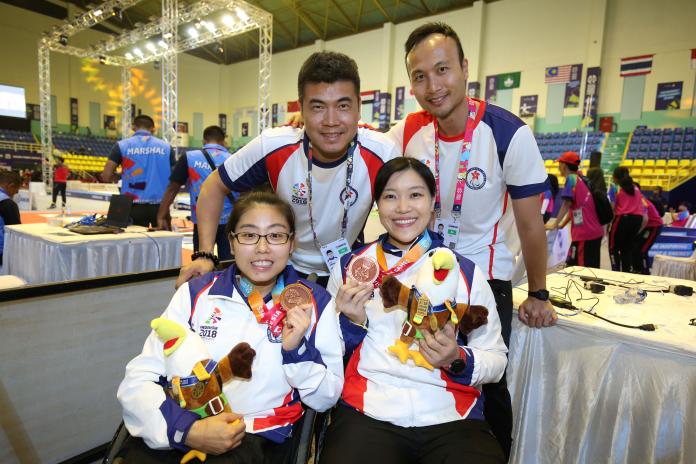 A team photo of two female athletes in wheelchairs, holding up bronze medals, and two coaches standing behind them.