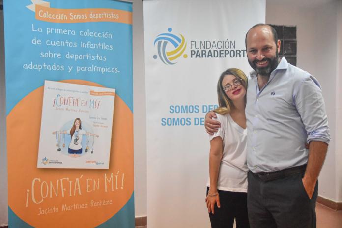 A man in a blue shirt has his arm over the shoulder of a young woman as they pose next to a poster announcing a new book release.