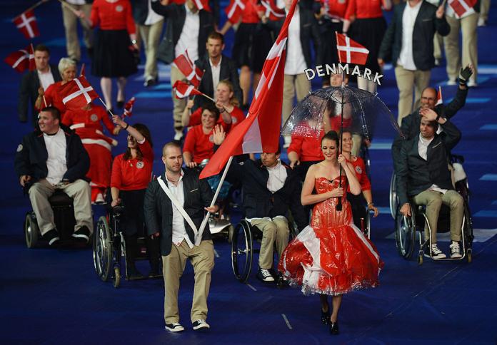 A male athlete carries the Danish flag in the Opening Ceremony of the London 2012 Paralympic Games. He leads a group of about 10 athletes and walks behind a women in a red dress holding an umbrella with the words Denmark. 