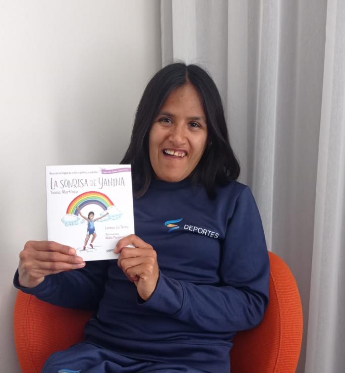 A female athlete in casual wear sits in an armchair as she smiles and holds up the book titled "La sonrisa de Yanina".