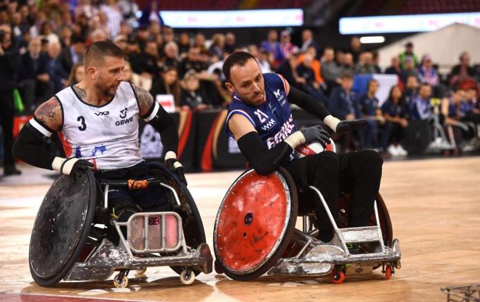 A male wheelchair rugby player carries the ball during a game, while another athlete watches the ball. 