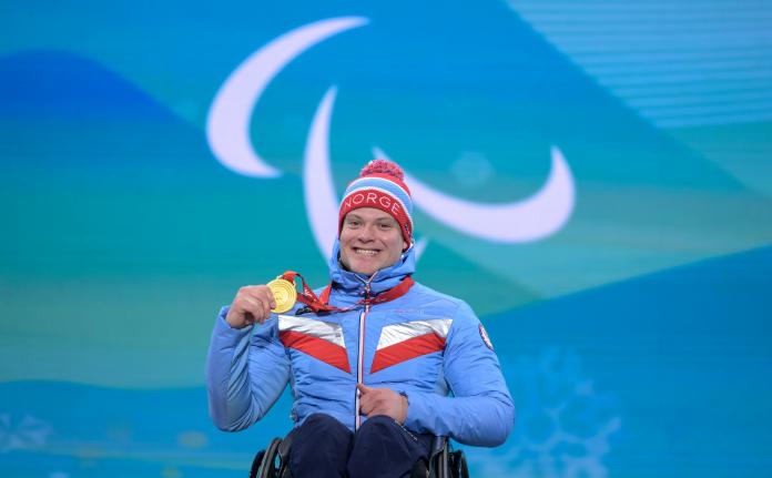 A male athlete holds a gold medal and smiles. The Three Agitos logo is in the background.