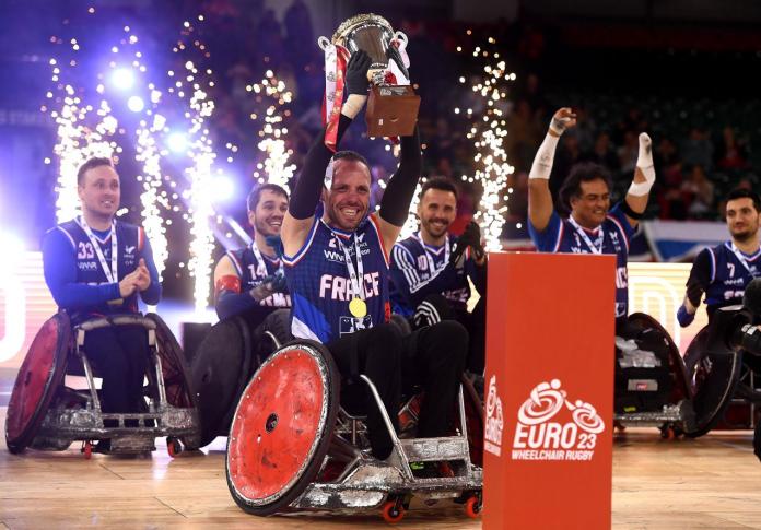 A male wheelchair rugby player lifts a trophy, while five teammates celebrate behind him.