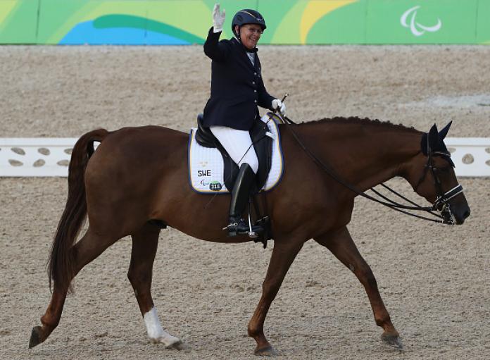 A female athlete waves her hand while riding a horse.