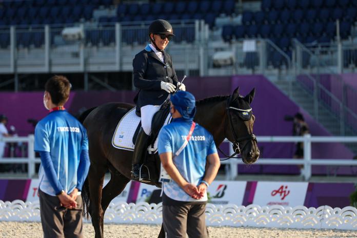 A female athlete rides a horse at the Tokyo 2020 Paralympic Games, while watched by two volunteers wearing blue shirts.