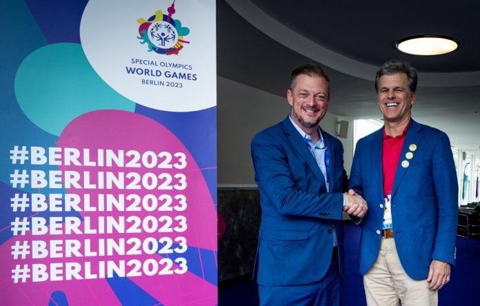 Two men shake hands next to a banner of the Special Olympics World Games Berlin 2023.