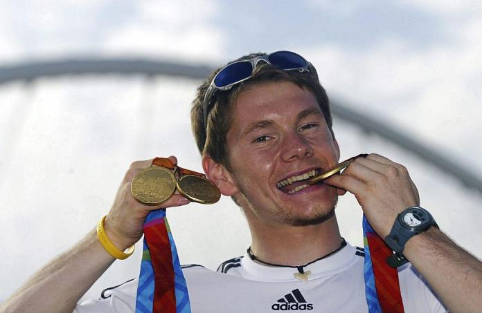 A male athlete poses for a photo by biting a gold medal and raises two other gold medals in his right hand.