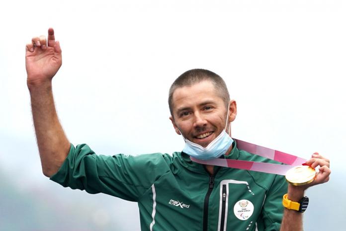 A male athlete raises his right arm in celebration while holding a gold medal with his left hand.