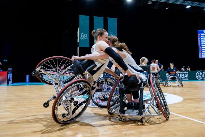 Two female wheelchair basketball players hug at an indoor gymnasium.