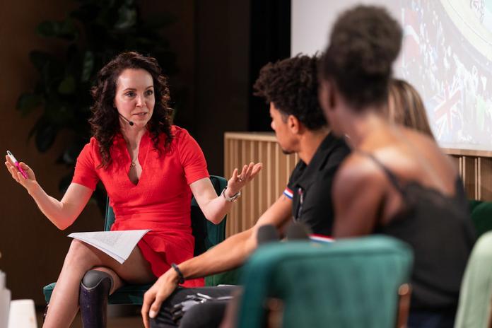 A female Paralympian moderates a session during the IPC summit. She is wearing a red dress and opens her arms as she speaks.