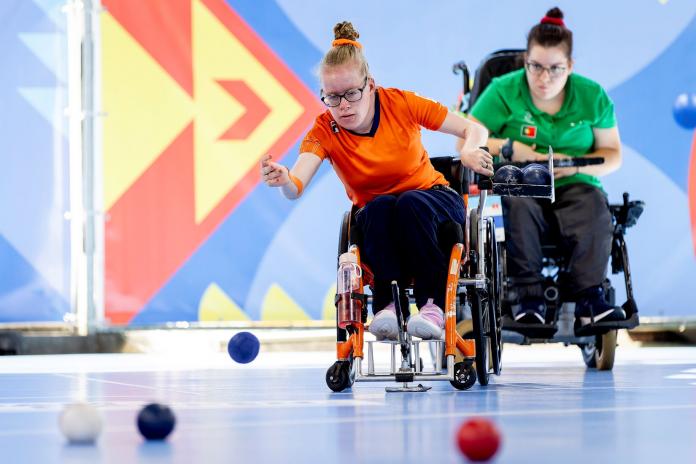 A female athlete throws a blue ball during a boccia match. Another female athlete is behind her.