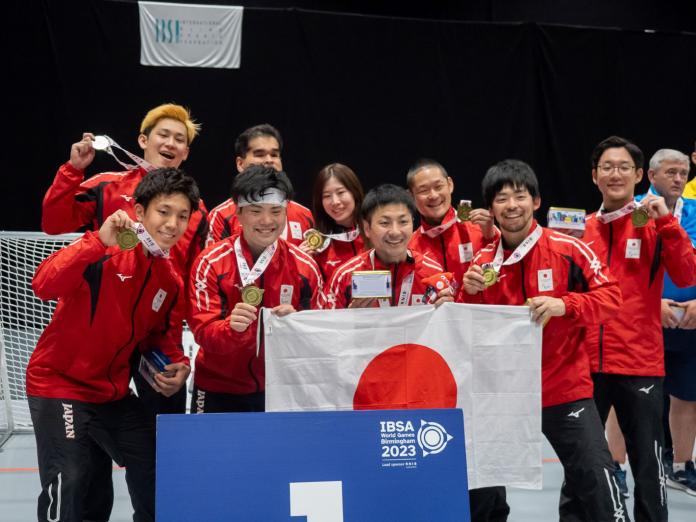 9人の選手とスタッフが日本の国旗を持って金メダルを持ってポーズを取っている。