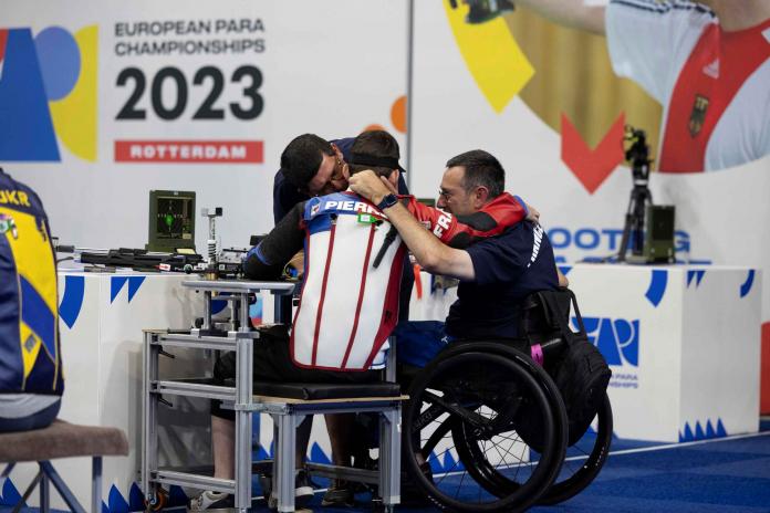 Tekerlekli sandalyedeki bir adam diğer iki adama sarılıyor