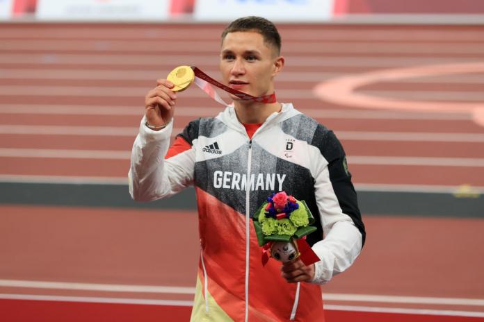 A male athlete holds a gold medal next to his face.