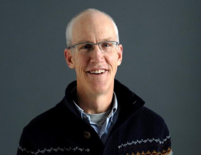 A man smiles in front of a gray background.