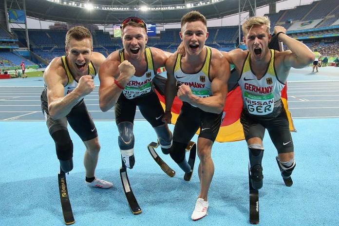 Four German male Para athletes wearing running blades celebrate after winning a race.
