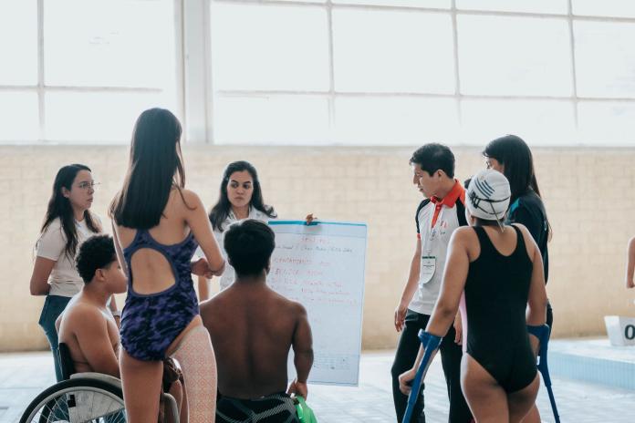 A woman holds up a board in front of seven people, including Para swimmers