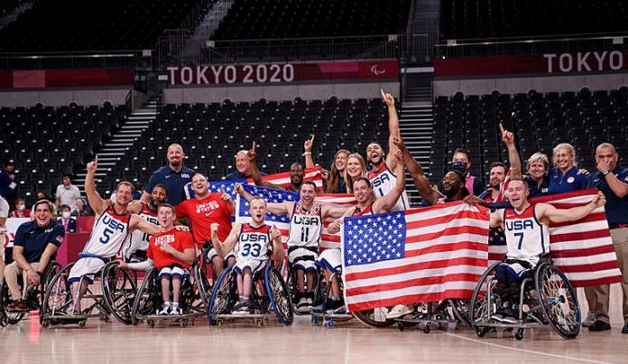 Athletes and staff of the USA wheelchair basketball team celebrate on the court. They pose for a photo and some of the athletes are holding an American flag.