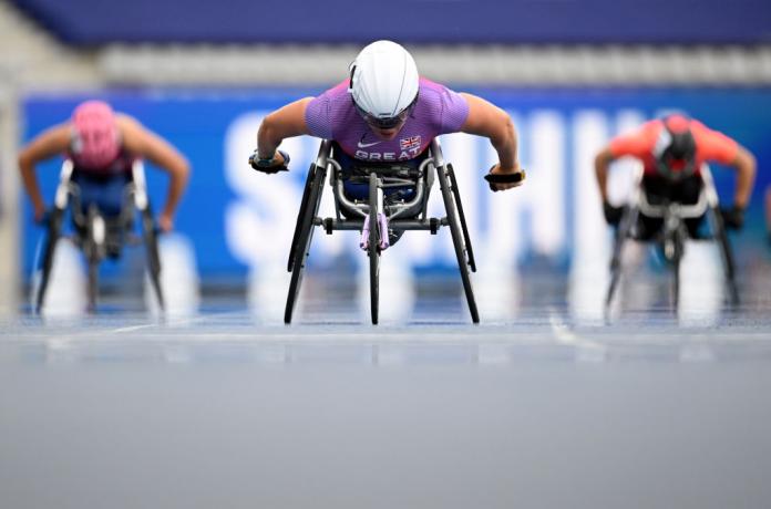 Three female wheelchair racers compete. The athlete in the centre is wearing Great Britain's team jersey, and leads against the other two athletes.