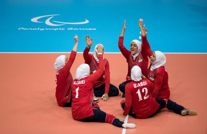 Six female athletes in red uniform celebrate on court at the Rio 2016 Paralympics. They form a circle and raise their hands