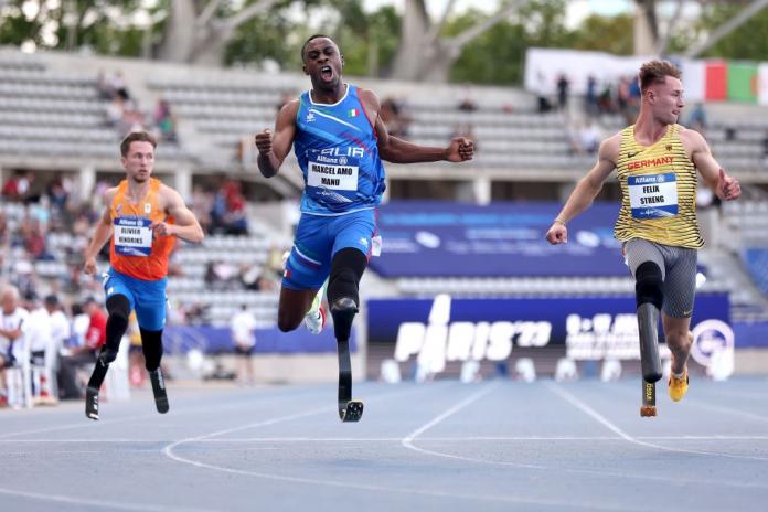 Three male athletes compete in a race. An Italian athlete in the centre pumps up his fists.