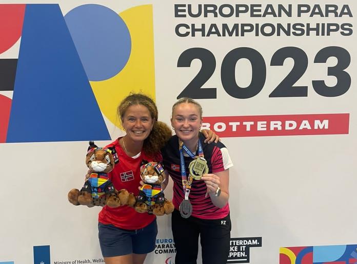 A female coach and a female Para badminton player. The athlete holds a gold medal while the coach holds two stuffed animals of a mascot.