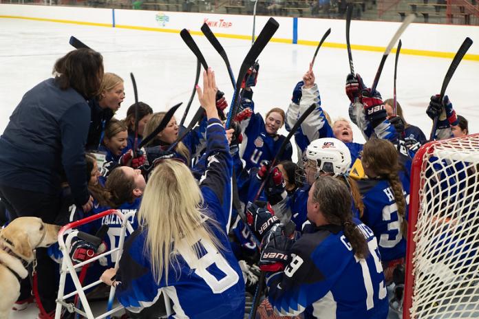 About 15 female Para ice hockey players celebrate on the ice by raising their sticks.