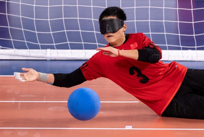 A male athlete wearing an eyeshade saves a ball in front of a goalball net.