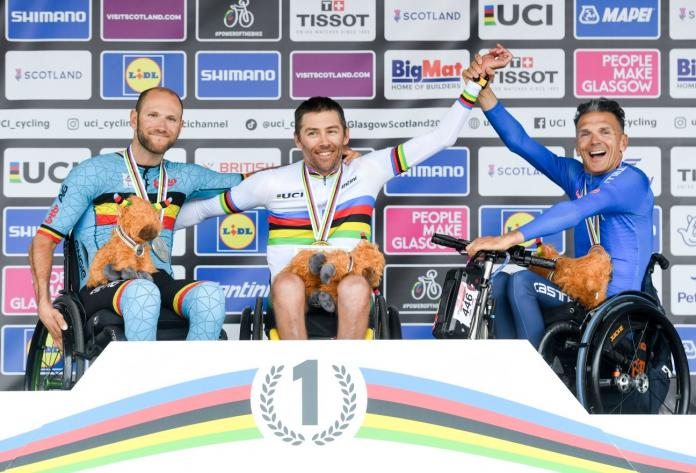 Three male athletes pose for a photo on a podium. The athlete in the centre is wearing the UCI rainbow jersey.