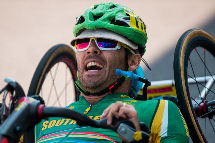 Tres atletas masculinos posan para una fotografía en un podio. El atleta del centro lleva el maillot arcoíris de la UCI.