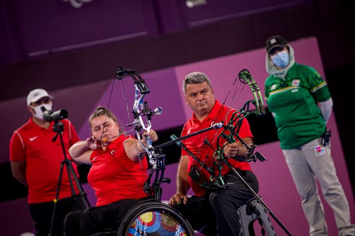 A female archer prepares to shoot an arrow. A male athlete is sitting next to her. 