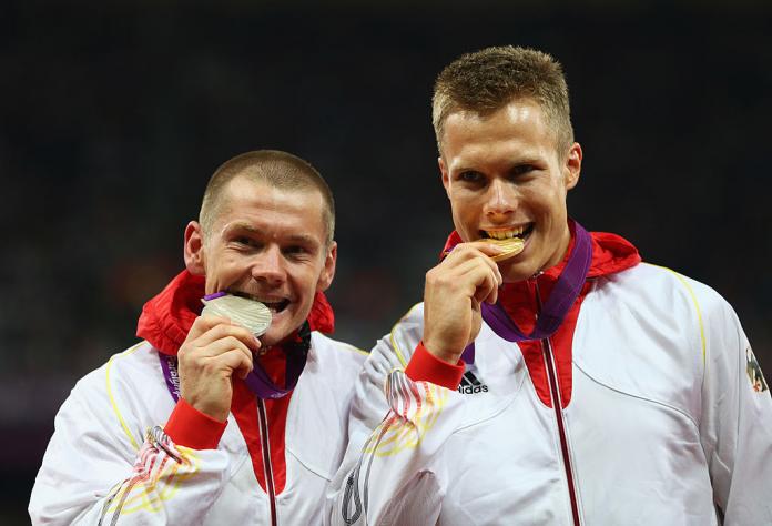 Two male athletes bite their medals. The athlete on the left is biting a silver medal, and the athlete on the right is biting a gold.