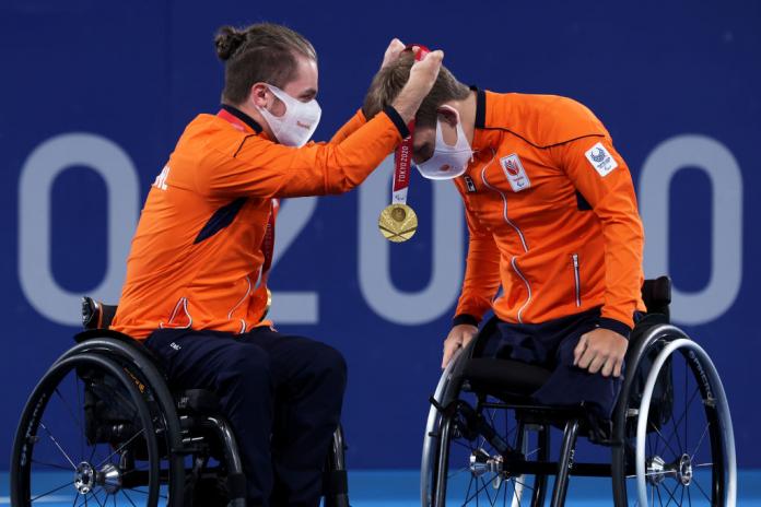 A male wheelchair tennis player presents his doubles partner with a gold medal.