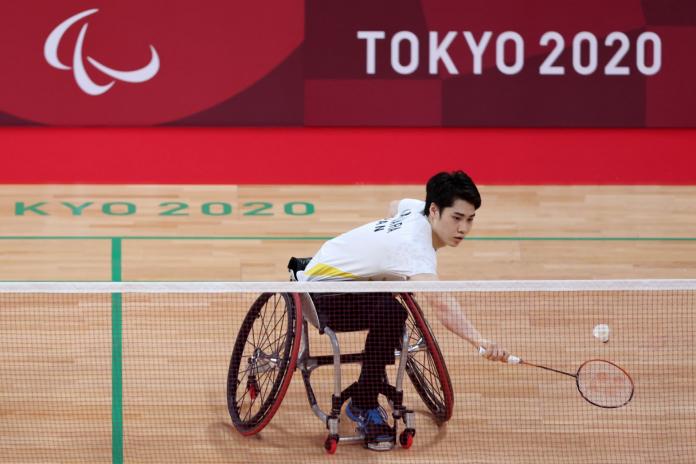 A male wheelchair badminton player reaches to return a shuttle during a match.