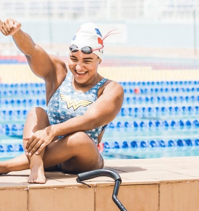 A female Para swimmer wearing a Wonder Woman swimsuit sitting by a pool.